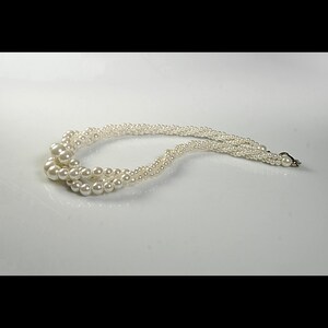May include: A cream-coloured, multi-strand pearl necklace. The necklace features varying sizes of round pearls, with a clasp closure. The strands are intertwined, creating a layered effect. The necklace is displayed on a white surface.