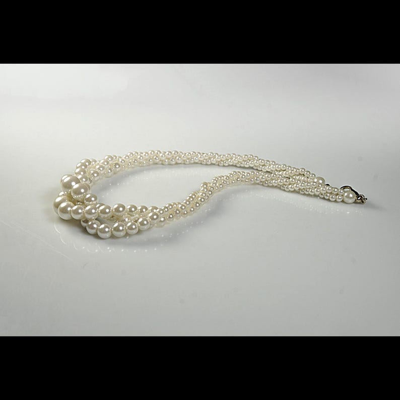 May include: A cream-coloured, multi-strand pearl necklace. The necklace features varying sizes of round pearls, with a clasp closure. The strands are intertwined, creating a layered effect. The necklace is displayed on a white surface.