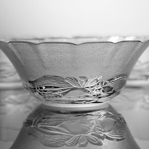 May include: A set of clear glass bowls with a textured surface and a decorative floral design around the base. The bowls have a scalloped rim and are reflected on a shiny surface.