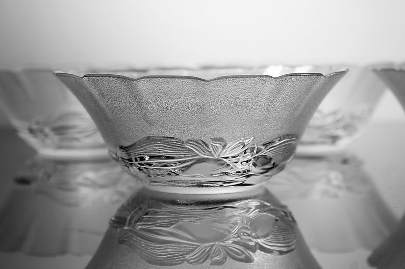 May include: A set of clear glass bowls with a textured surface and a decorative floral design around the base. The bowls have a scalloped rim and are reflected on a shiny surface.