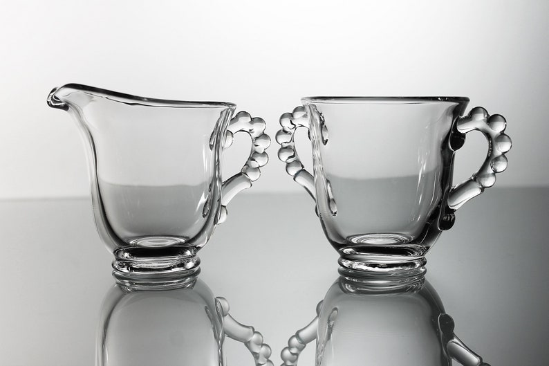 K&ouml;nnte beinhalten: Ein Set aus zwei klaren Glaskr&uuml;gen f&uuml;r Sahne und Zucker mit perlenbesetzten Griffen. Die Kr&uuml;ge sind leer und stehen auf einer reflektierenden Oberfl&auml;che.