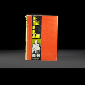Puede incluir: Un libro de tapa dura rojo titulado "The Trial of Dr. Adams" de Sybille Bedford, autora de "A Legacy". La portada presenta una foto en blanco y negro de un hombre sosteniendo un periódico.