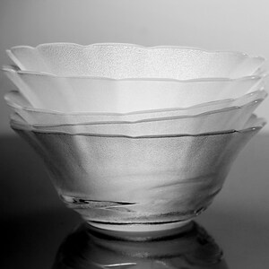 May include: A stack of four clear glass bowls with a textured, frosted appearance. The bowls have a scalloped rim and are stacked on top of each other, creating a layered effect. The bowls are reflected on a dark surface.