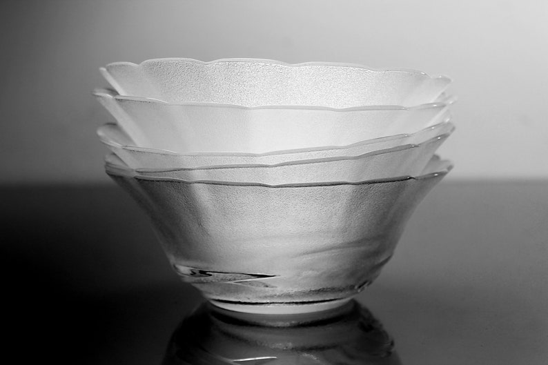 May include: A stack of four clear glass bowls with a textured, frosted appearance. The bowls have a scalloped rim and are stacked on top of each other, creating a layered effect. The bowls are reflected on a dark surface.