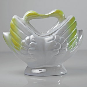 May include: White ceramic swan-shaped planter with a green and yellow glaze. The planter has a handle and is designed to hold small plants or flowers.