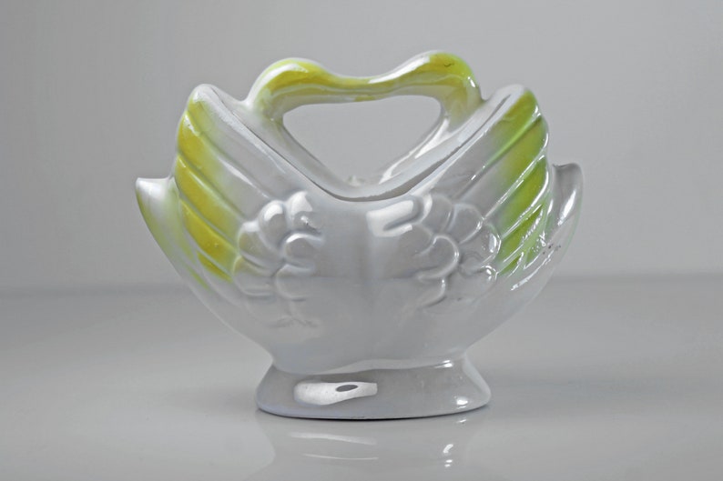 May include: White ceramic swan-shaped planter with a green and yellow glaze. The planter has a handle and is designed to hold small plants or flowers.