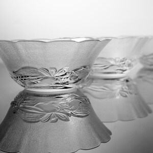 May include: Three clear glass bowls with a frosted texture and a floral design around the base. The bowls have a scalloped rim and are arranged in a row, reflecting on a shiny surface.
