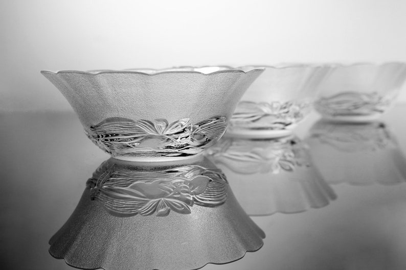 May include: Three clear glass bowls with a frosted texture and a floral design around the base. The bowls have a scalloped rim and are arranged in a row, reflecting on a shiny surface.