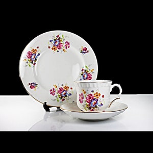 Puede incluir: Un juego de taza y platillo de té blanco con un patrón floral en tonos de rosa, azul y amarillo. El juego tiene un borde dorado.