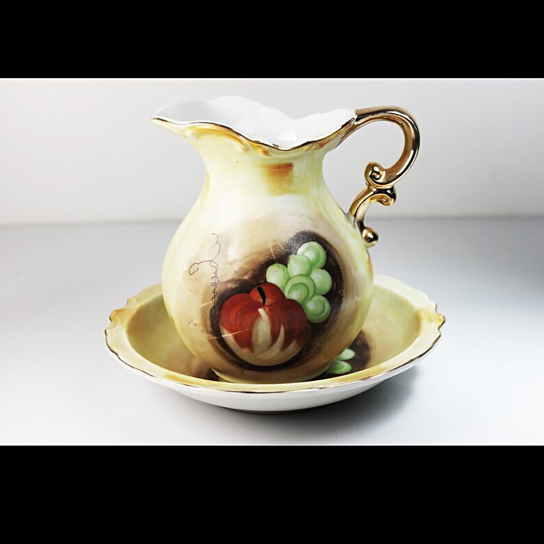 Puede incluir: Un juego vintage de jarra y cuenco de porcelana con un dise&ntilde;o pintado a mano de fruta, incluyendo una manzana y uvas. La jarra tiene un asa dorada y el juego tiene un esquema de color amarillo y marr&oacute;n.