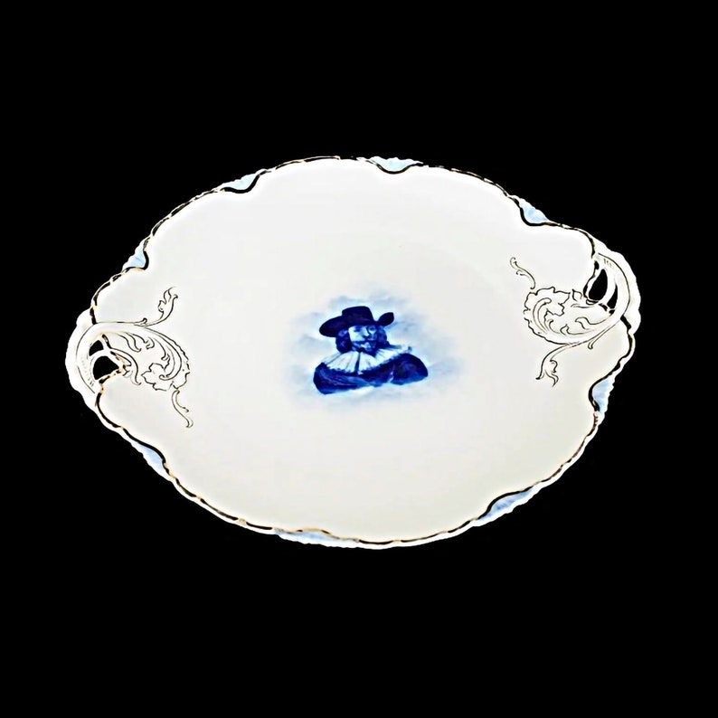May include: A white porcelain serving platter with a blue and white image of a man in a hat. The platter has gold trim and two ornate handles.