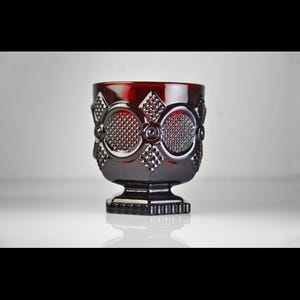 May include: A ruby red glass goblet with a detailed pattern. The goblet features a square base and a textured design of circles, diamonds, and other geometric shapes. The glass has a glossy finish and reflects light.