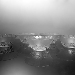 May include: Five frosted glass bowls with scalloped edges are arranged on a reflective surface. The bowls have a floral design on the base. The image is in black and white, highlighting the texture and shape of the bowls.