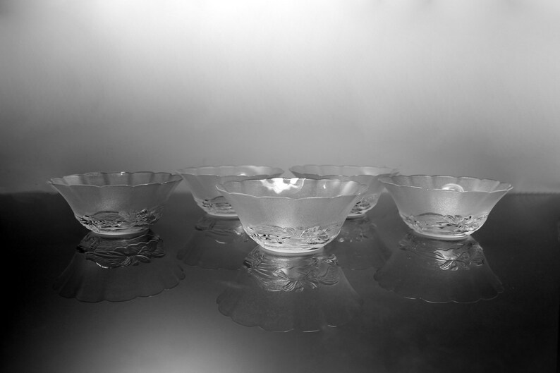 May include: Five frosted glass bowls with scalloped edges are arranged on a reflective surface. The bowls have a floral design on the base. The image is in black and white, highlighting the texture and shape of the bowls.