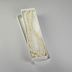 May include: A cream pearl necklace in a white rectangular box. The necklace features varying pearl sizes and a gold clasp. The box is open, revealing a white fabric lining. The pearls are cream-colored.