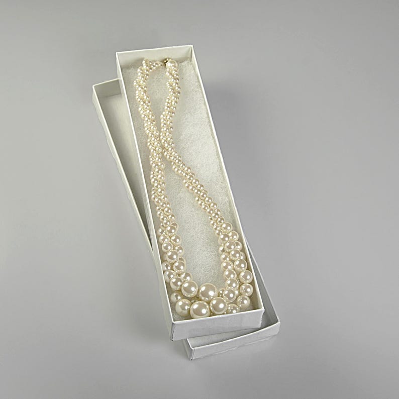 May include: A cream pearl necklace in a white rectangular box. The necklace features varying pearl sizes and a gold clasp. The box is open, revealing a white fabric lining. The pearls are cream-colored.