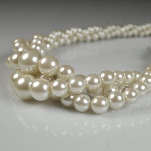 May include: Close-up of a strand of faux pearls. The pearls are a creamy white colour and vary in size, with larger pearls in the foreground. The pearls are strung together, creating a necklace or bracelet.