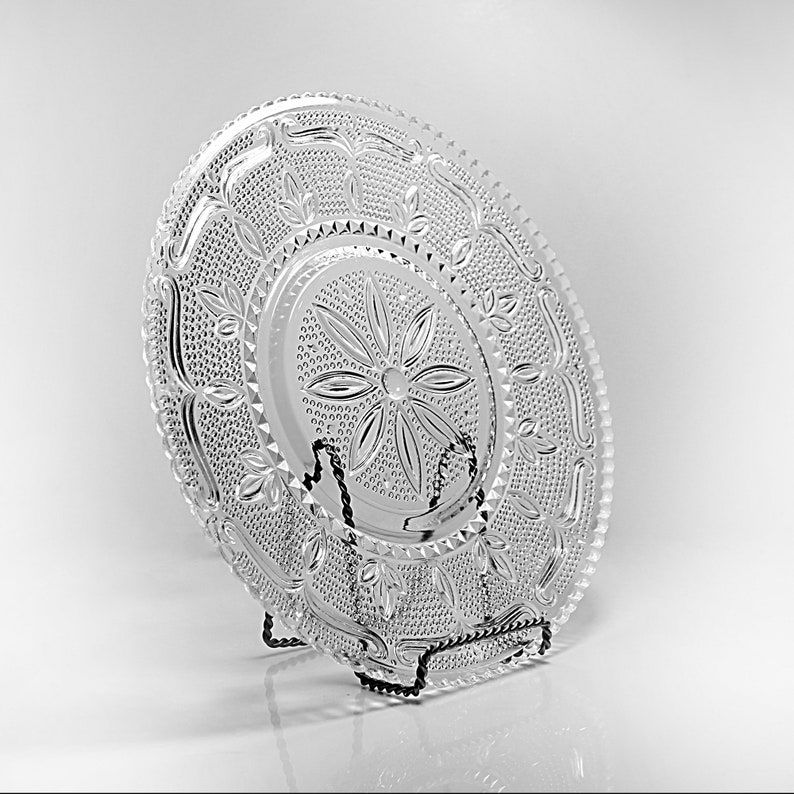 Op de afbeelding: Een heldere glazen serveerschaal met een decoratief bloemmotief. De schaal staat op een zwarte metalen standaard.