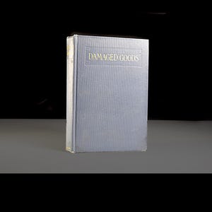 May include: A blue-grey hardcover book with the title "DAMAGED GOODS" in gold lettering. The book is standing upright, with the spine visible. The edges of the book show signs of wear. The background is dark.