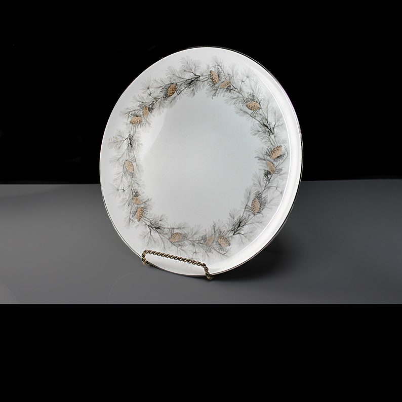 Op de afbeelding: Een witte keramische serveerschaal met een gouden rand en een ontwerp van zwarte en gouden dennenappels.