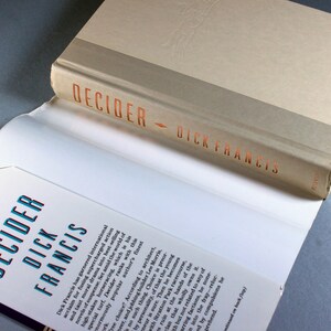 May include: A close-up of the spine of a paperback book titled "Decider" by Dick Francis. The book cover is white with a brown band at the top. The title is printed in orange letters.