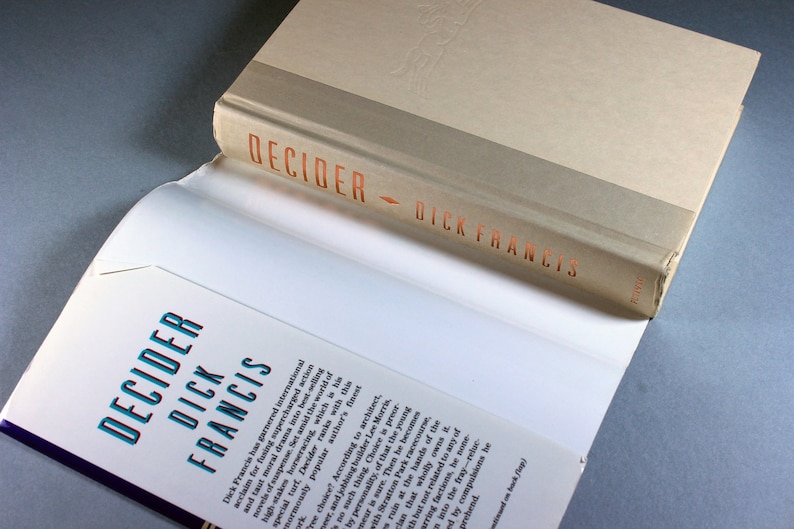 May include: A close-up of the spine of a paperback book titled "Decider" by Dick Francis. The book cover is white with a brown band at the top. The title is printed in orange letters.