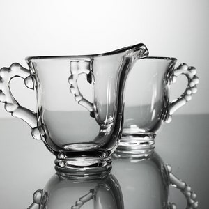 K&ouml;nnte beinhalten: Zwei klare Glask&auml;nnchen mit herzf&ouml;rmigen Griffen. Die K&auml;nnchen sind aus Glas und haben eine glatte, polierte Oberfl&auml;che.