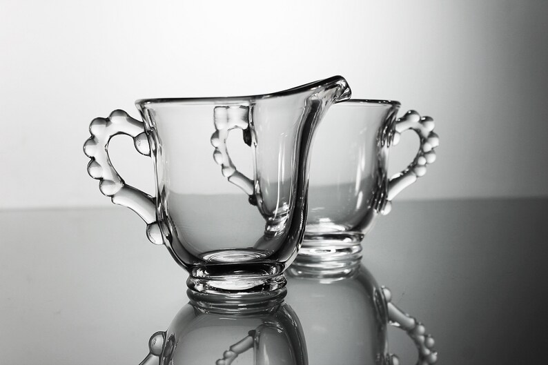 K&ouml;nnte beinhalten: Zwei klare Glask&auml;nnchen mit herzf&ouml;rmigen Griffen. Die K&auml;nnchen sind aus Glas und haben eine glatte, polierte Oberfl&auml;che.