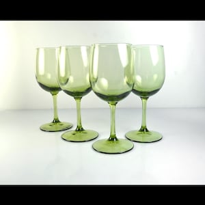 May include: Four green-tinted wine glasses are arranged on a white surface. The glasses have a classic wine glass shape with a rounded bowl and a long stem. The color of the glass is a pale green.