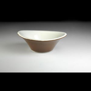 May include: A small, brown ceramic bowl with a white interior. The bowl has a unique, curved shape and a smooth, glossy finish. The rim is slightly flared, and the bowl appears to be empty. The background is a gradient of white to black.