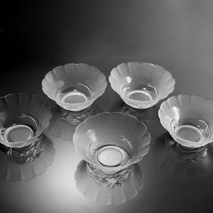 May include: Five clear glass bowls with a scalloped rim. The bowls are arranged in a circular pattern, with their reflections visible below. The bowls have a frosted appearance and a decorative design at the base.