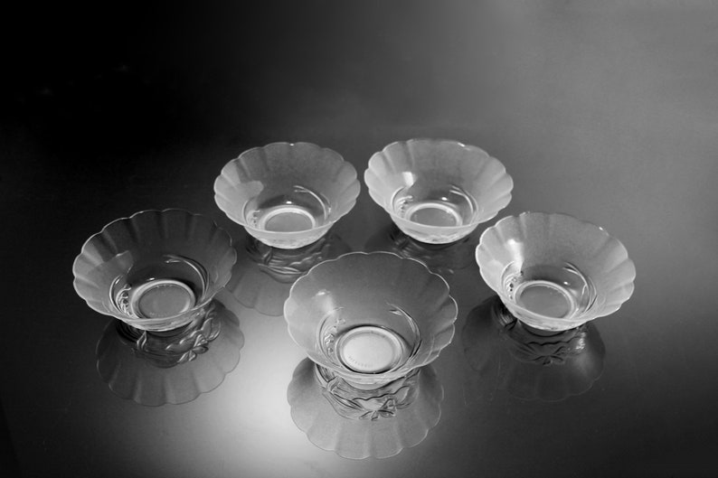 May include: Five clear glass bowls with a scalloped rim. The bowls are arranged in a circular pattern, with their reflections visible below. The bowls have a frosted appearance and a decorative design at the base.