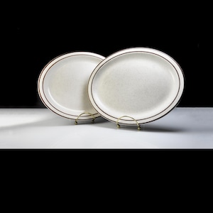May include: Two oval ceramic plates with a speckled off-white surface and a brown border. The plates are displayed on small, gold-coloured plate stands against a black background. The plates are likely for serving food.