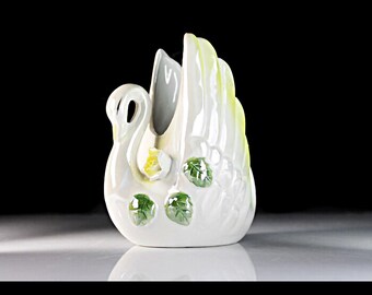 May include: White ceramic swan napkin holder with green leaves and a yellow flower.