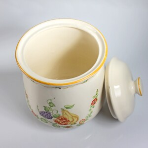 May include: A white ceramic canister with a floral design and a matching lid. The canister has a yellow rim and a floral pattern with green leaves, red roses, purple grapes, and a yellow pear.