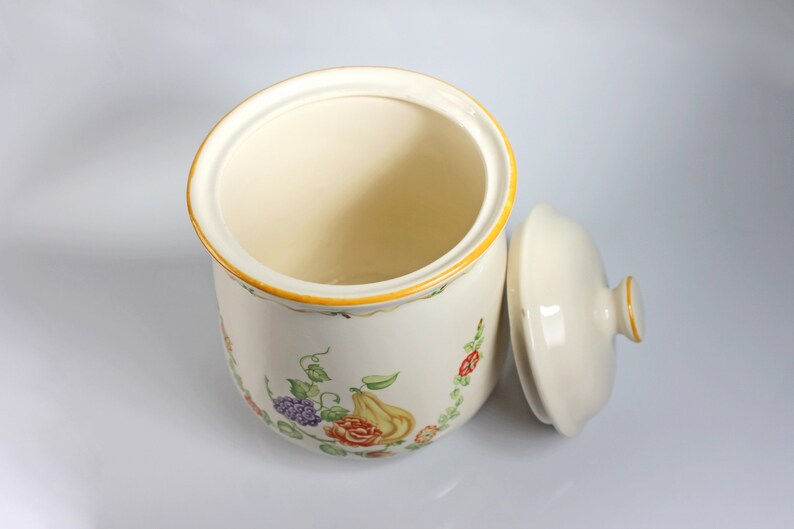 May include: A white ceramic canister with a floral design and a matching lid. The canister has a yellow rim and a floral pattern with green leaves, red roses, purple grapes, and a yellow pear.