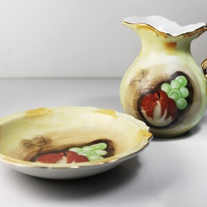 Puede incluir: Un juego de dos platos de porcelana vintage con un dise&ntilde;o pintado a mano de fruta, incluyendo una manzana y uvas. Los platos son amarillos con acentos marrones y verdes. El juego incluye una jarra peque&ntilde;a y un taz&oacute;n.