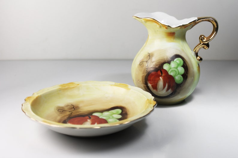 Puede incluir: Un juego de dos platos de porcelana vintage con un dise&ntilde;o pintado a mano de fruta, incluyendo una manzana y uvas. Los platos son amarillos con acentos marrones y verdes. El juego incluye una jarra peque&ntilde;a y un taz&oacute;n.