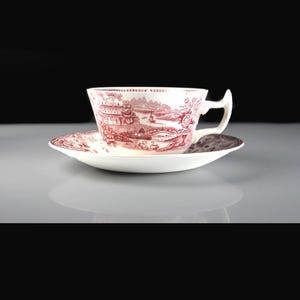 May include: A white teacup and saucer set with a red and white landscape design. The teacup has a curved handle and sits on a matching saucer. The design features buildings, trees, and water elements.
