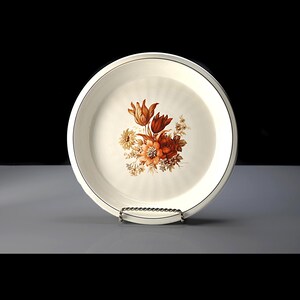 May include: A cream-coloured ceramic plate featuring a floral design in orange, red, and yellow hues. The plate has a thin black border and is displayed on a silver metal stand. The floral arrangement is centrally positioned on the plate.