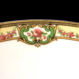 May include: Close-up of the rim of a white porcelain plate with a gold and green floral design. The design features pink roses and green leaves.