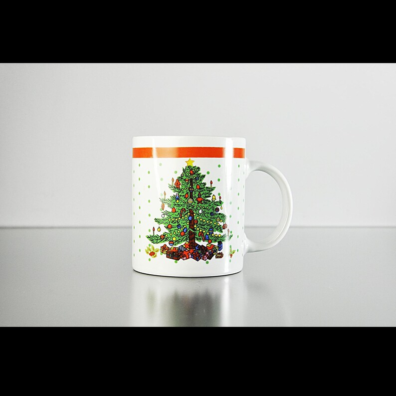 May include: White ceramic mug featuring a green Christmas tree design. The tree is adorned with red, blue and yellow baubles and has a yellow star on top. The mug has a red and white polka dot pattern and a red stripe around the top.