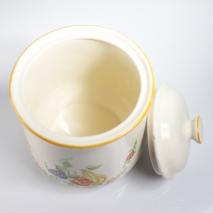 May include: A white ceramic canister with a floral design and a matching lid. The canister has a yellow rim and features a floral pattern with red, yellow, and purple flowers.