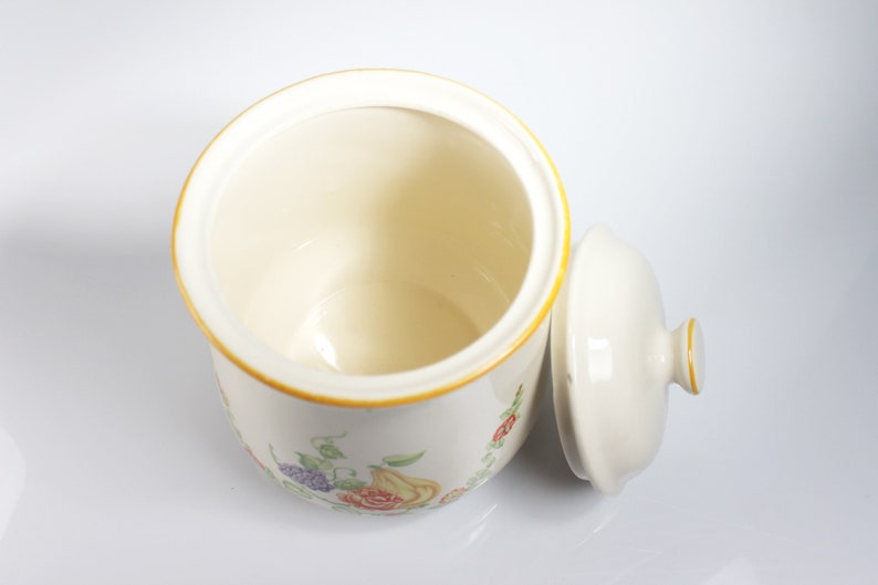 May include: A white ceramic canister with a floral design and a matching lid. The canister has a yellow rim and features a floral pattern with red, yellow, and purple flowers.