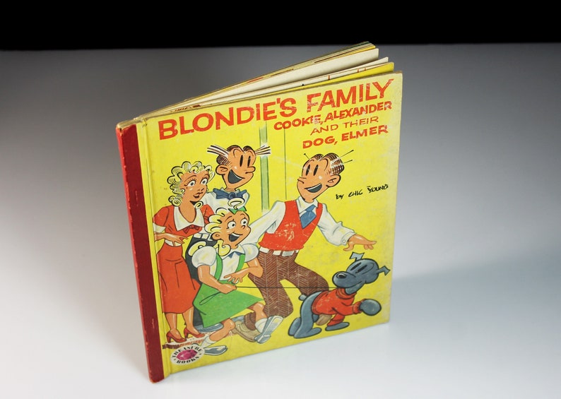 May include: A yellow book cover with the title "Blondie's Family Cookie, Alexander and Their Dog, Elmer" by Chic Young. The cover features a cartoon illustration of Blondie, Dagwood, Alexander, and Elmer.