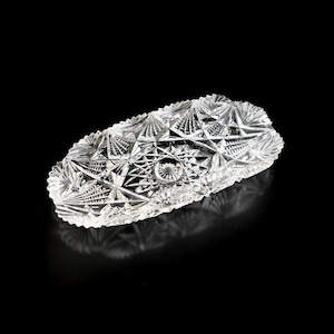 May include: A clear, oval glass dish with detailed starburst and geometric patterns. The dish has a scalloped edge, designed to reflect light. The symmetrical design is suitable for serving or display. The dish measures approximately 20 cm in length.