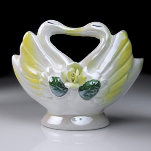 May include: White ceramic swan vase with yellow and green accents. The swans are facing each other with their necks forming a heart shape. A yellow rose is in the center of the vase.