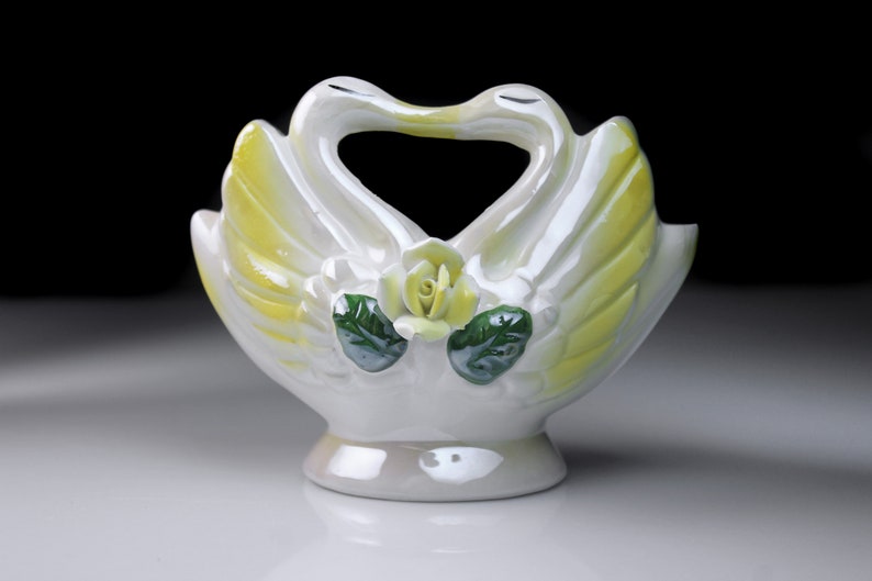 May include: White ceramic swan vase with yellow and green accents. The swans are facing each other with their necks forming a heart shape. A yellow rose is in the center of the vase.