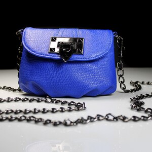 Pode incluir: Uma bolsa transversal azul cobalto com acabamento texturizado. A bolsa apresenta um fecho prateado e uma al&ccedil;a de corrente. A bolsa est&aacute; sobre uma superf&iacute;cie branca com um fundo preto.