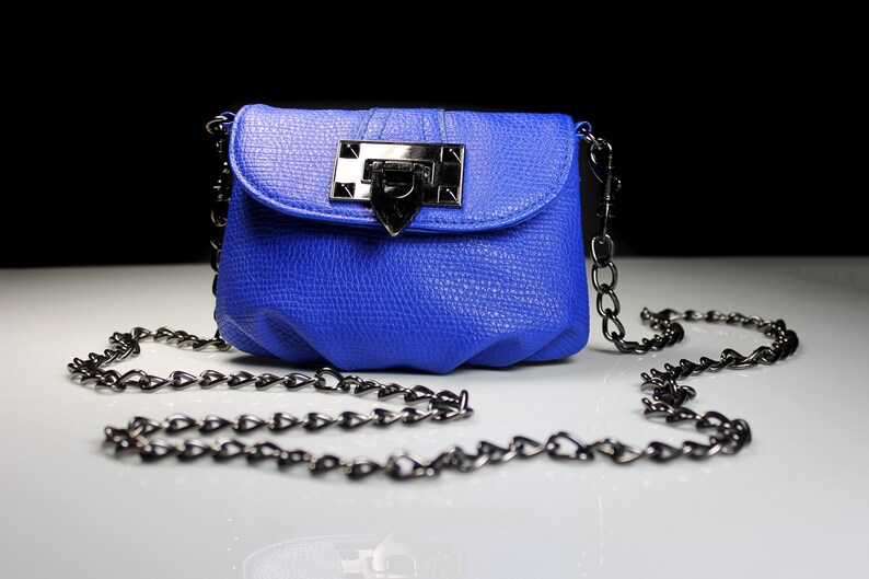 Pode incluir: Uma bolsa transversal azul cobalto com acabamento texturizado. A bolsa apresenta um fecho prateado e uma al&ccedil;a de corrente. A bolsa est&aacute; sobre uma superf&iacute;cie branca com um fundo preto.